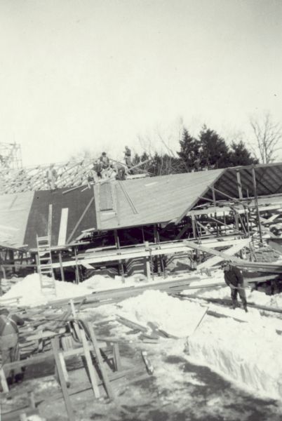 Elevated view of construction workers near a truss on the roof. In the foreground on the right another man is working around piles of snow and building materials on the ground.