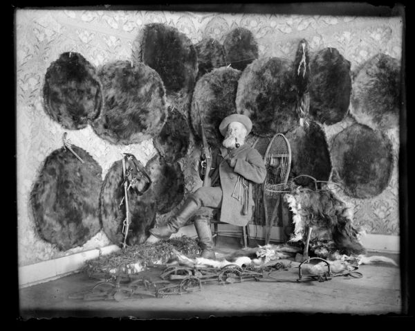 Lewis Adam Struble is seated in the corner of a room with a number of beaver pelts displayed on the walls behind him. He is sitting in a chair wearing a coat, hat and boots, and is holding a rifle and smoking a pipe. Also on display are guns, belts, snowshoes, knives, traps, pelts, and an axe. A taxidermied snake is displayed on the floor beside him in a box.