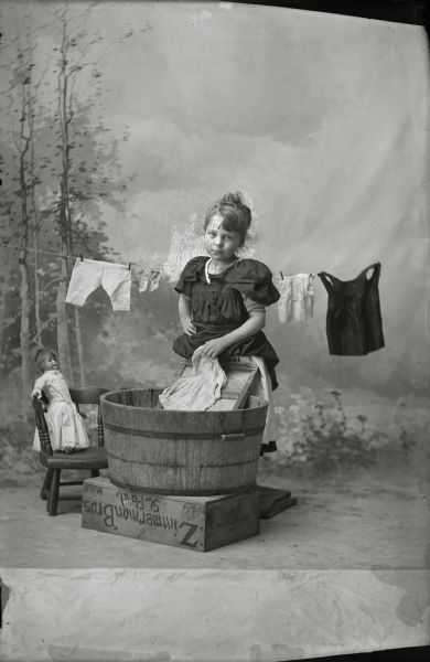 Studio portrait in front of a painted backdrop of Bertha Gesell washing doll clothes in a wooden tub. Clothes are on a clothesline behind her. Her doll sits in a chair on the left.