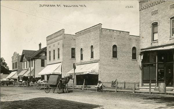 View across Jefferson Street toward several storefronts and horse-drawn vehicles on the right. Near the Gustave [__]ner Harness Shop is a man seated on a wheelbarrow near the sidewalk. Next door on the left is a meat market. The brick building on the right is a General Store/Post Office. Caption reads: "Jefferson Street, Sullivan, Wis."
