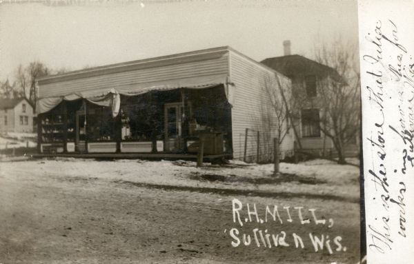 View across snowy, unpaved street towards the storefront. Caption reads: "R.H. Mill, Sullivan, Wis."