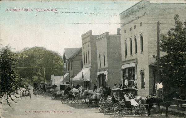 Partially colorized postcard view down Jefferson Street. People are gathered in front of one of the storefronts on the right, and horse-drawn vehicles are parked along the curb. Caption reads: "Jefferson Street, Sullivan, Wis."