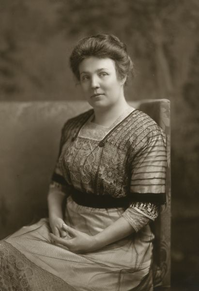 Studio portrait of Meta Berger seated with her hands folded in her lap. The dress was made at the Milwaukee Girls' Trade School (founded in 1909 by Lizzie Kander) in 1910 for Mrs. Berger to wear in Washington, D.C., when she accompanied her husband there for his first term as a US congressman. The dress in this photograph is located in the collections of the Wisconsin Historical Museum (1946.912).