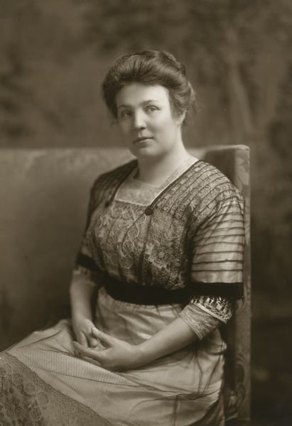 Studio portrait of Meta Berger seated with her hands folded in her lap. The dress was made at the Milwaukee Girls' Trade School (founded in 1909 by Lizzie Kander) in 1910 for Mrs. Berger to wear in Washington, D.C., when she accompanied her husband there for his first term as a US congressman. 

The dress in this photograph is located in the collections of the Wisconsin Historical Museum (1946.912).