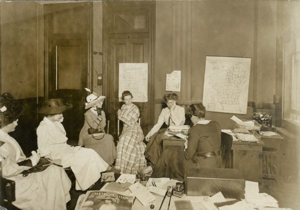 Group of women including Ada James (in checked dress at center) at the suffrage headquarters. A map of Wisconsin is hanging on the wall at right and another map, possibly Minnesota, hangs behind Ms. James. A calendar on the wall indicates June of 1918.