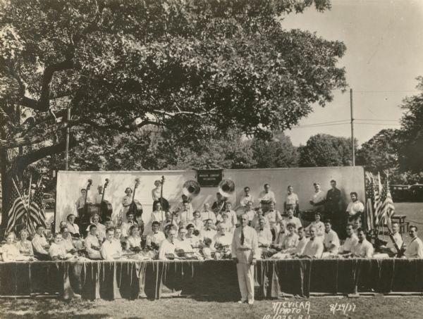 Madison Concert Orchestra members with their instruments seated on a "stage" under trees at Vilas Park. The conductor is standing in front. Groups of U.S. flags are displayed at the sides of the stage.