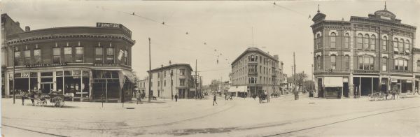 Panoramic view of the downtown towards Bascom Hall from the corner of the Capitol Square, showing Carroll, Mifflin, and State Streets. Several businesses can be seen, including: A. Haswell & Company Furniture Company, J.P. Meuer Labor Hall, Dr. S.J. Fryette Osteopathic Physician, Kioscke Brothers Hardware, Menges Pharmacy, and Savings Loan & Trust Company. There are signs for many other business along State Street but they are not legible. Pedestrians are walking, pulling carts, and driving horse-drawn carriages. A streetcar is on the 200 block of State Street. The streets are lined with power lines and street lights are suspended over intersections.