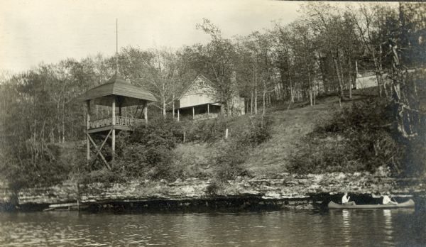 View from the water (probably Lake Mendota) of summer cottages at Blackhawk. There is a pavilion at the shoreline. There are two people canoeing near the shore.