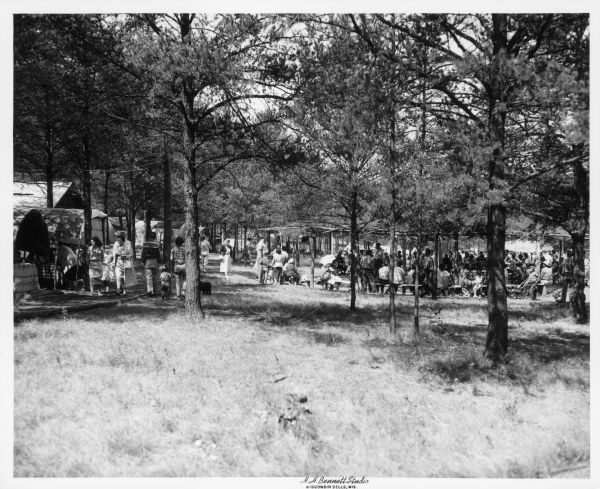 View across grass towards a group of Ho-Chunk people sitting together at their summertime village, which was managed by Roland "Pipe" Dyer. There are trees in the foreground and behind the village.