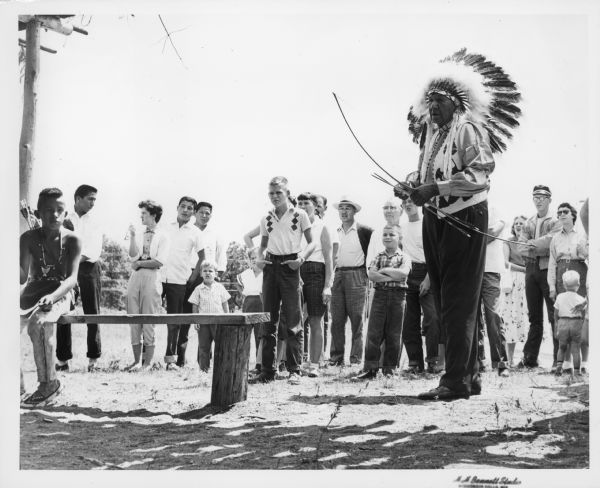 Sam Blowsnake prepares to shoot an arrow with his bow as a crowd of people watches behind him.