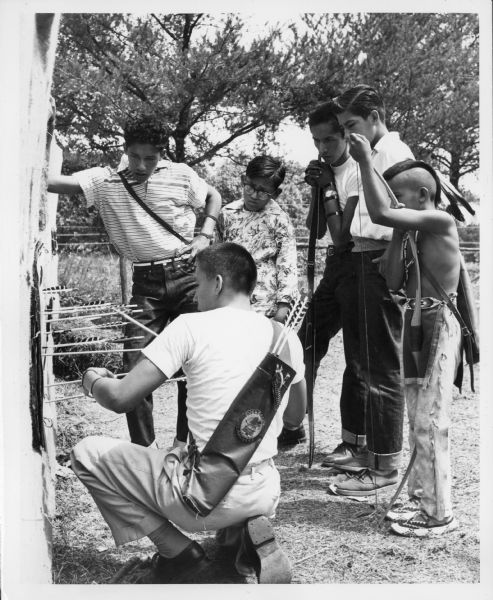 Six Native American boys and teenagers take part in an archery competition. They are holding their bows and carrying arrows in a bag on their backs. Riley Sine is calling off scores to the scorekeeper.