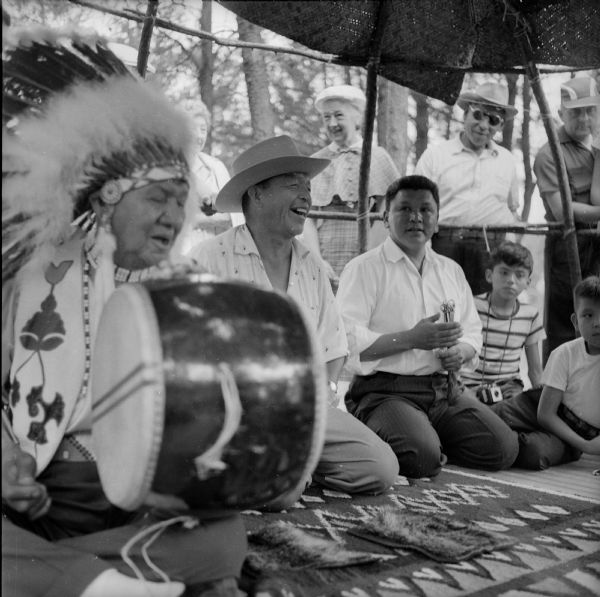 Several Indian men kneel on a rug to play the moccasin game. One man is holding a drum and wearing a headdress (also called a war bonnet).