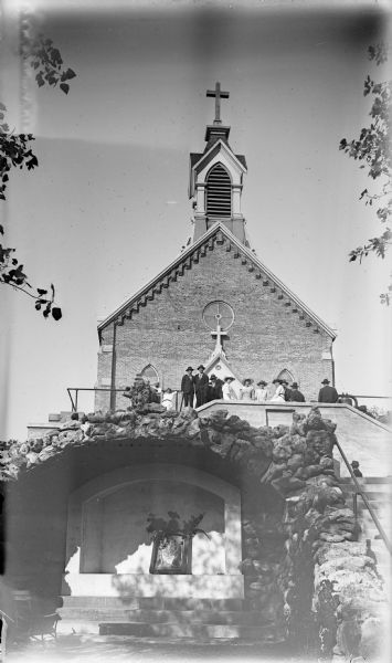 Exterior view of a group of people at Holy Hill. They are standing above a shrine. There is a large bell beside the group on the right.