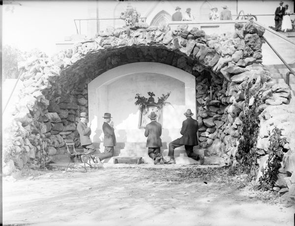 Four men kneeling and praying at the shrine at Holy Hill. More people are gathered near the church above them.
