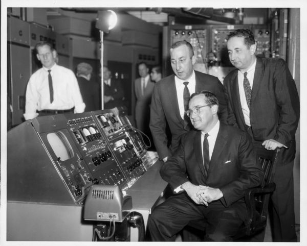 Newton Minow is seated at a monitor in a broadcasting control room. Two men stand behind him, looking at the monitor over his shoulder. Other people are in the background on the left.