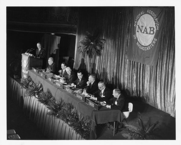 Elevated view of Newton Minow (seated fourth from the right) attending a NAB conference. He is sitting at a table on a stage with six other men. Another man stands at a podium on the far left.