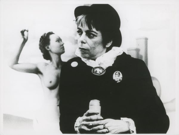 Waist-up seated portrait of Shirley Clarke. She is holding a microphone and wearing a bowler hat and a black outfit with a white shirt, as well as a "Portrait of Jason" button. In the background is a painting or mural of a nude woman.