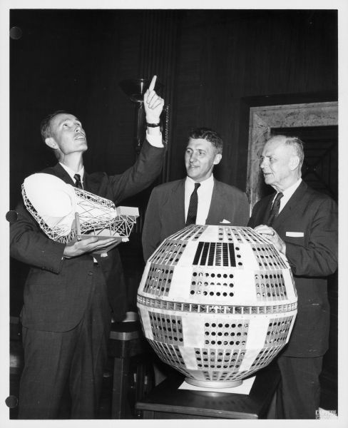 Members of the Senate Commerce Communications Subcommittee stand with a model of the Telstar satellite. A man holds a piece of the model and is pointing skyward.
