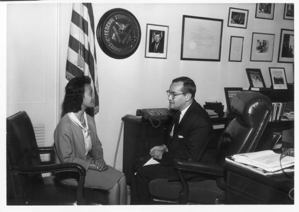 Newton Minow and a woman on his staff are facing each other, having a discussion. There is a portrait of John F. Kennedy hanging on the wall behind them.