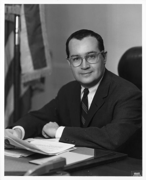 Formal waist-up portrait of Newton Minow at his desk, holding papers.