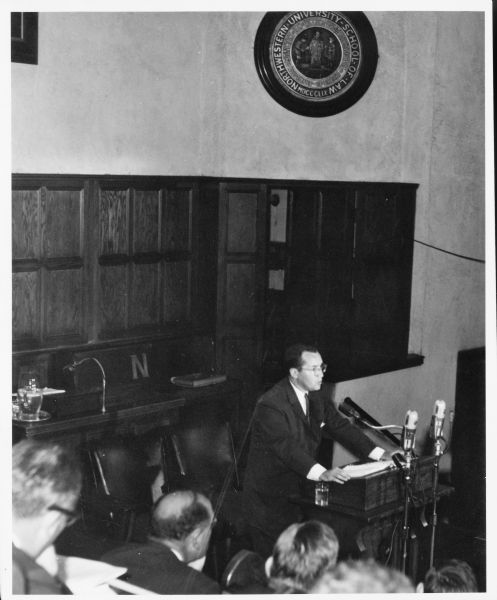 Slightly elevated view over audience towards Newton Minow giving a speech while standing at a podium. A large plaque on the wall behind him says "Northwestern University School of Law."
