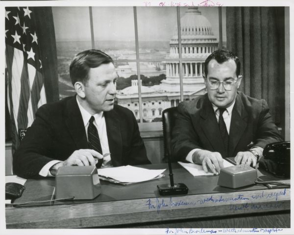 John Brodemas (sp?) and Newton Minow are seated at a desk together, which has a microphone and a telephone. Behind them is a fake window with a view of the U.S. Capitol and an American Flag.