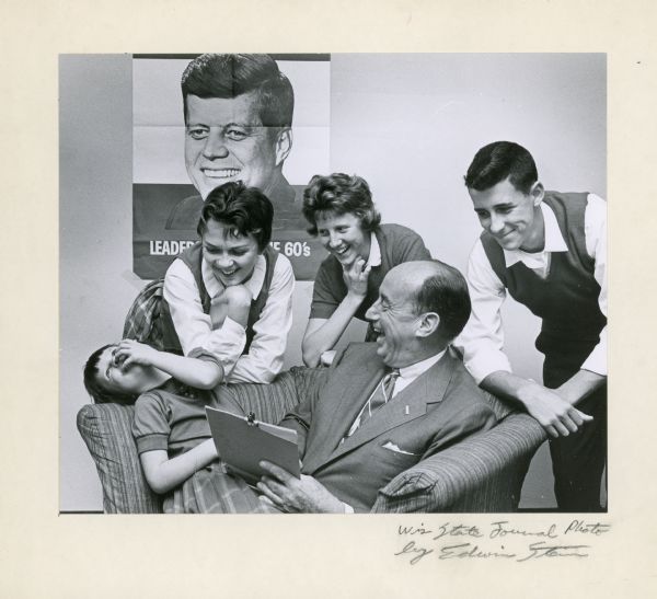 Adlai Stevenson, seated, laughs with the children of Jim Oogle. There is a poster of John F. Kennedy on the walll in the background.