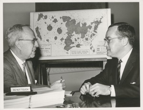 Newton Minow and another man are talking while seated in front of a map on the wall.