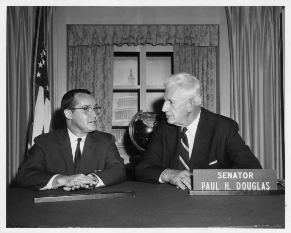Newton Minow and Senator Paul H. Douglas are seated side-by-side at a desk. Behind them is a globe in front of a fake curtained window with a view of the capitol building, and an American flag on the left, with the stage set U.S. Capitol flanked by stage curtains.