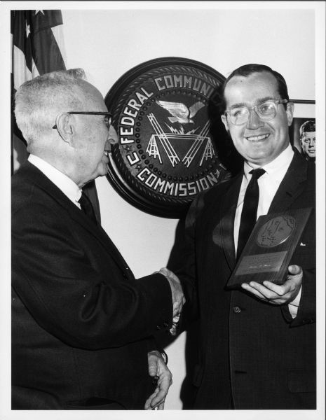 Newton Minow and Leo Solomon shake hands in front of a Federal Communications Commission (FCC) plaque. Minow appears to be receiving an award.