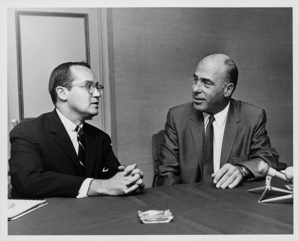 Newton Minow is seated at a table with another man with a microphone on the table.