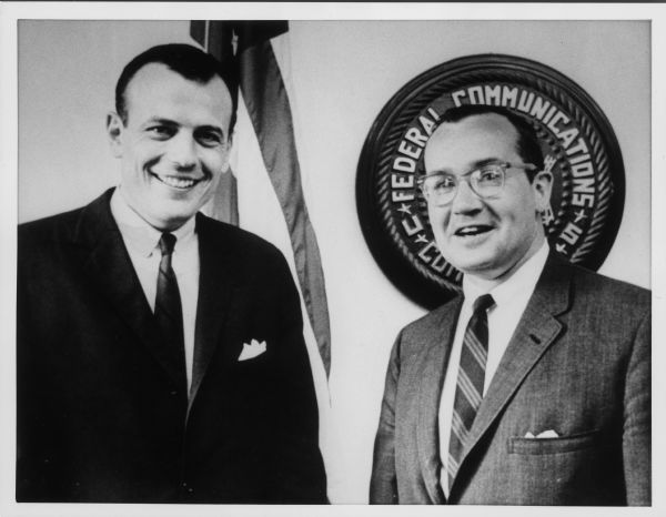 Newton Minow and Bill Henry stand together in front of an American Flag and a Federal Communications Commission (FCC) plaque.