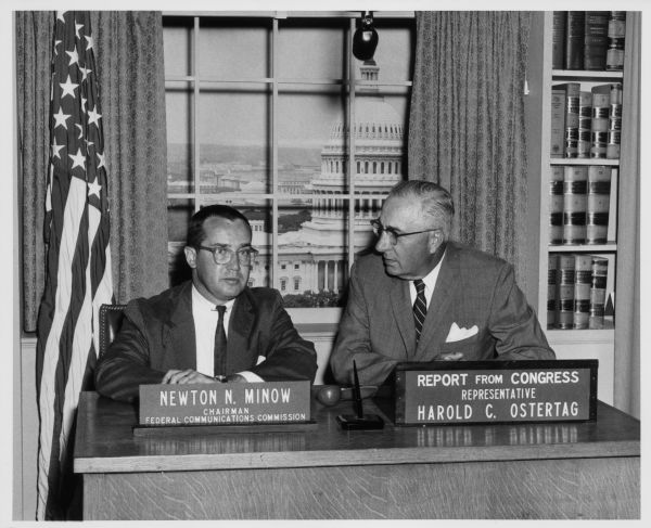 Newton Minow, chairman of the Federal Communications Comission (FCC), and Harold C. Ostertag, Report From Congress representative are seated together with a microphone above them. Behind them on a stage set is a fake window with a view of the U.S. Capitol, books in a bookcase, and an American Flag.