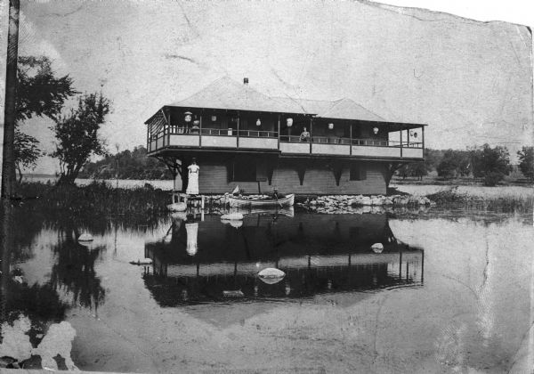 View across water towards Robert Lamp's cottage, Rocky Roost, which was designed by Frank Lloyd Wright. Lamp's four nieces are posed on the balcony, on the pier and in a canoe. The cottage is decorated with lanterns for a party.