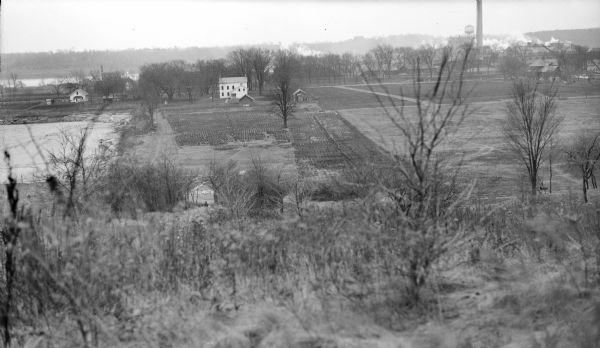 View from hill looking down across fields at the property of Charles Grignon. Grignon Mansion in the background among trees, and beyond is the Fox River. On the far right in the background is a water tower and a smokestack near industrial buildings.