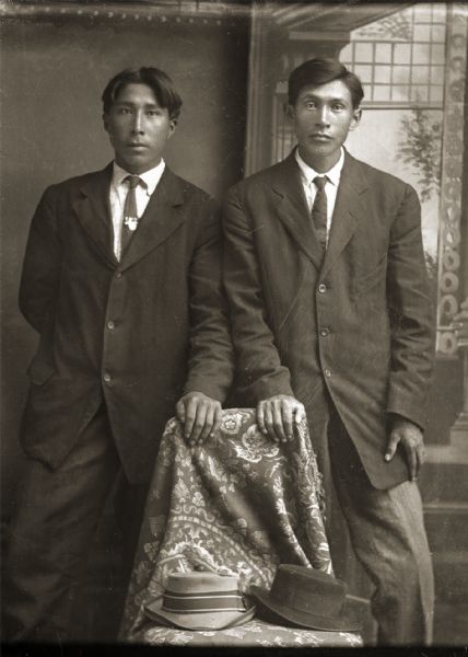 Studio portrait of two Ho-Chunk men posing standing in front of a painted backdrop. They have their hands resting on a draped chair holding two hats. Both men are wearing suits and neckties.