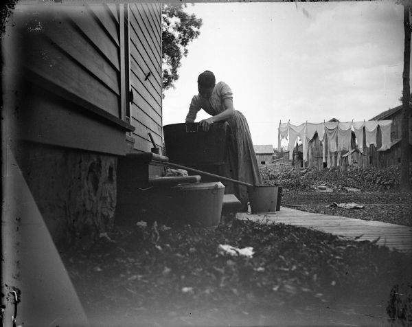 A woman washes clothes in a tub on the back steps of a farmhouse. In the background, clothes hang on a clothesline.