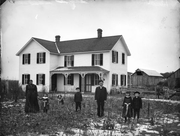 Group portrait of a man, woman, two boys, two girls, and a dog posing standing in the yard in front of a frame house and farm buildings.