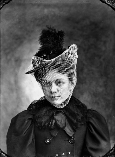 Studio portrait of a seated young woman wearing a hat with a veil and feather, and a lace collar over a puffy-sleeved jacket. Possibly Mrs. Cumnock.