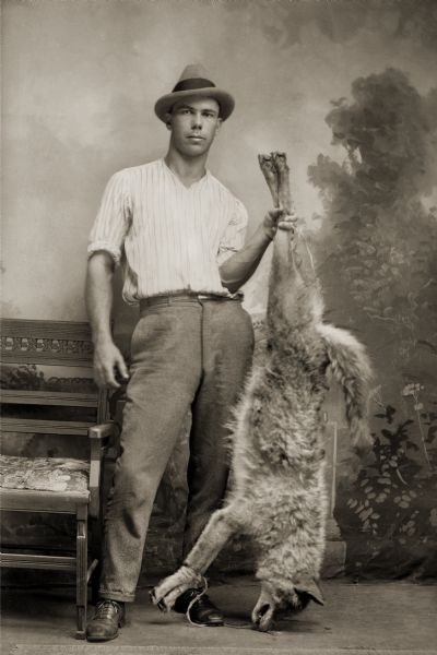 A man holds a dead wolf by the hindquarters and poses for a studio photograph in front of a painted backdrop. He is wearing a hat, shirt, and trousers.
