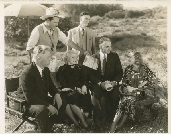 Outdoor group portrait taken on location for the film "Our Daily Bread." Barbara Pepper and Karen Morley are seated along with two men in director's chairs. King Vidor and another man stand behind them.  Vidor holds out a copy of the magazine "The Pacific Cooperator."