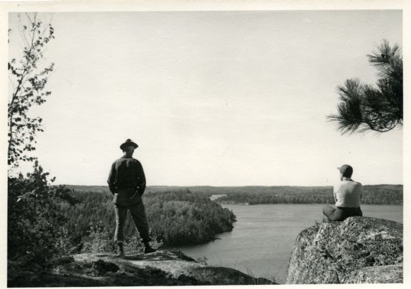 Sigurd F. Olson standing on cliff overlooking Robinson Lake in Quetico Provincial Park. Another person is sitting on a boulder next to where he stands.