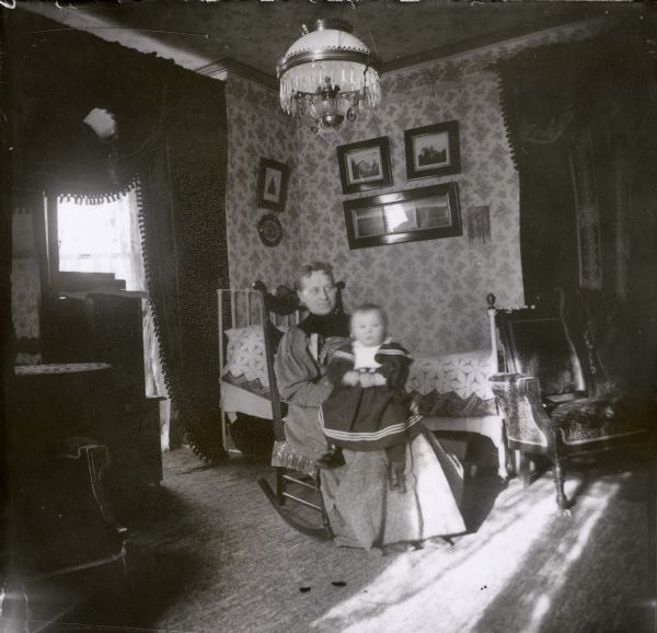 Indoor portrait of a woman seated in a rocking chair holding a baby on her knee. The infant is wearing a sailor dress.