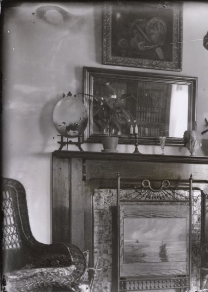 Interior view of a wicker chair near a fireplace with a decorative firescreen. There are candles and pieces of artwork on the mantle, and a mirror and painting hang on the wall.