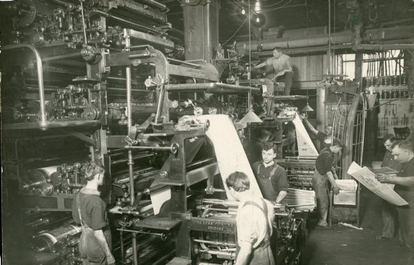 Several men work on a large printing press at the <i>Milwaukee Sentinel</i>. A man is stacking newspapers and two men are reading copies of the paper.