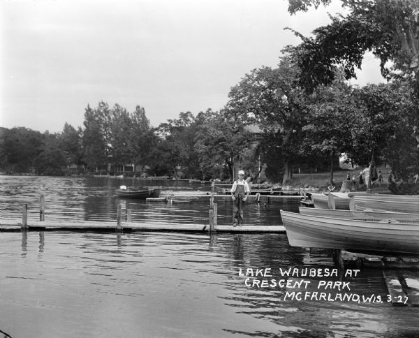 View across water towards a boy standing on a pier on Lake Waubesa. There are boats docked at the water's edge and a group of children and adults are on the shoreline in the background.