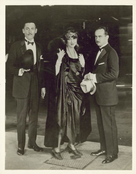 Lew Cody, Barbara La Marr, and Paul Bern attend the premiere of Norma Talmadge's film "Secrets" in Hollywood. The men are wearing tuxedos and Barbara wears a fancy dress and fur coat.