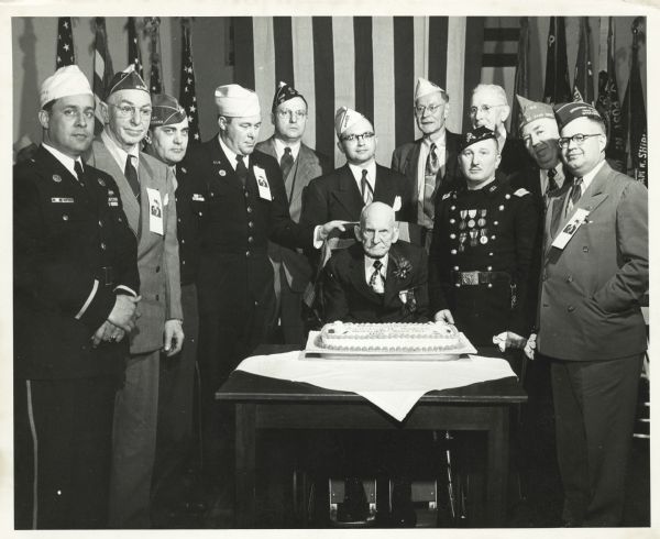 Group portrait of Lansing A. Wilcox — last Civil War veteran in Wisconsin — with a group of men at his 105th birthday. The birthday party was held at the Grand Army of the Republic home in King (now the Wisconsin Veterans Home). Mr. Wilcox is seated at a table behind a cake and surrounded by 11 unidentified men. Flags are displayed in the background.