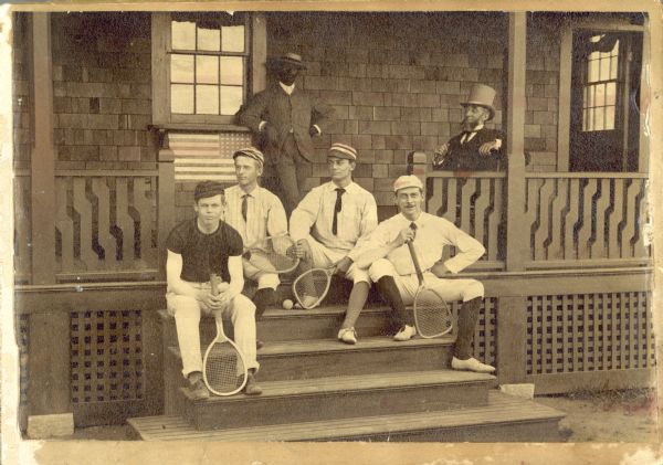 Charles Kendall Adams, president of the University of Wisconsin from 1892 to 1902, posing on a porch with five students. Below the window on the porch is an American Flag, in reverse. Four students are seated on the porch steps holding tennis rackets, and a fifth student is leaning against the wall; none of the men are identified.