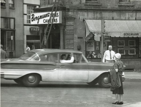 Photograph of a 1958 Chevy driving past Bergmann Prescription and Photo Center, located at 102 King Street. A man and a woman stand on opposite street corners watching the car pass through the intersection.